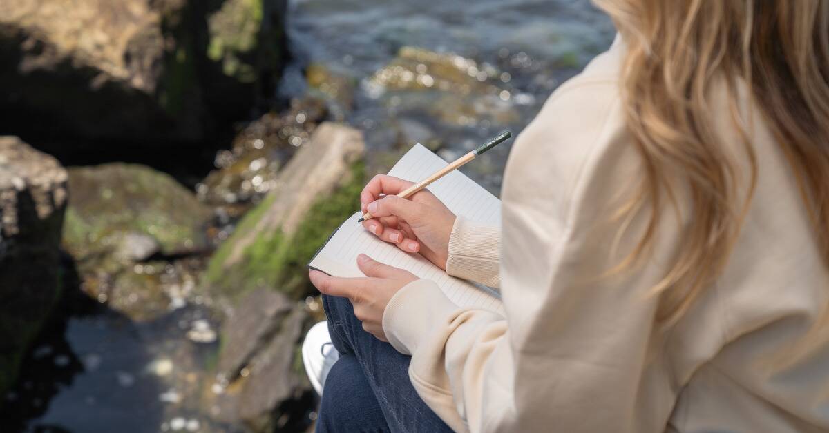 A woman writing in a small journal as she sits by some water.