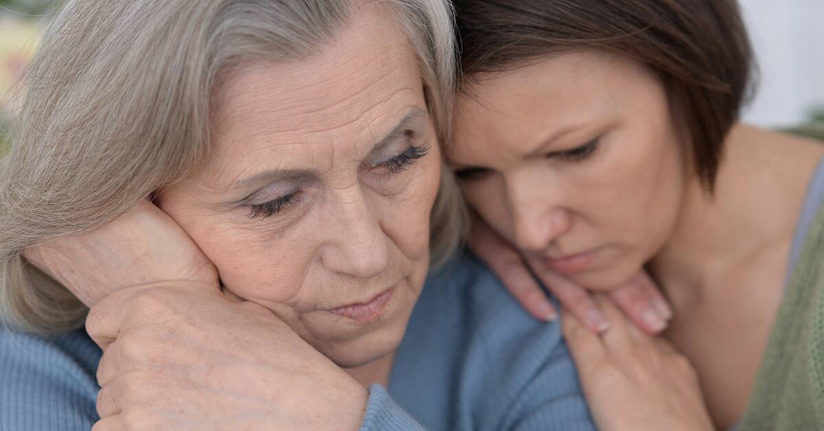 A close shot of a daughter leaning on her mother's shoulder, both looking sad.