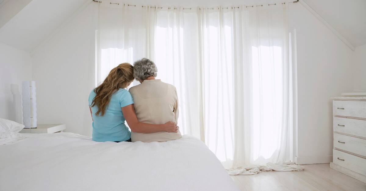 A mother and daughter sitting on a bed, daughter leaning into her mom and putting an arm around her.