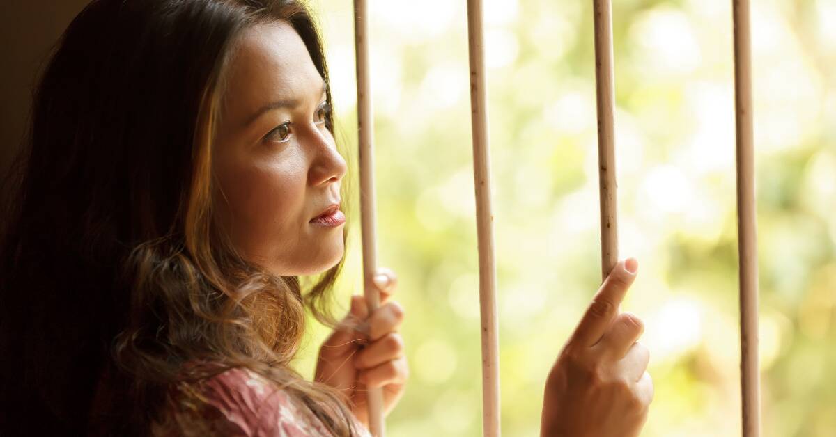 A woman looking forlornly out a barred window, holding onto the bars.