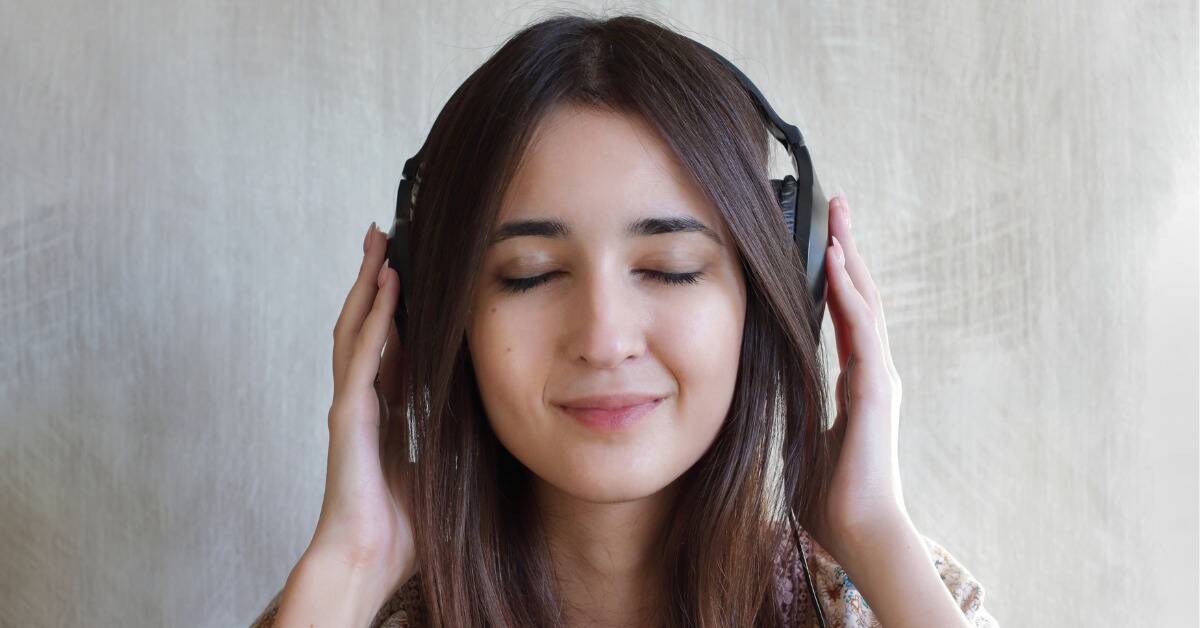 A woman smiling peacefully, wearing headphones, her hands gently resting on those headphones.