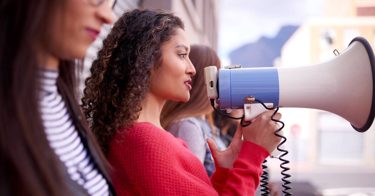 A line of women standing against a wall, one holding up a megaphone to her mouth.