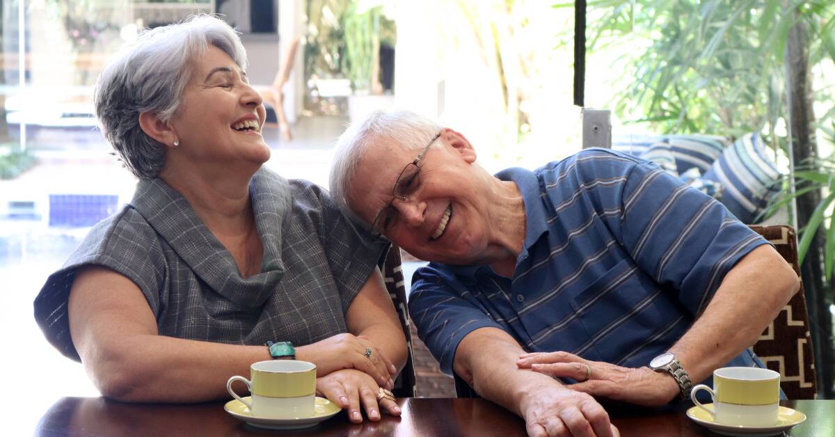 An older couple out for tea, both laughing as the man leans into the woman's shoulder.