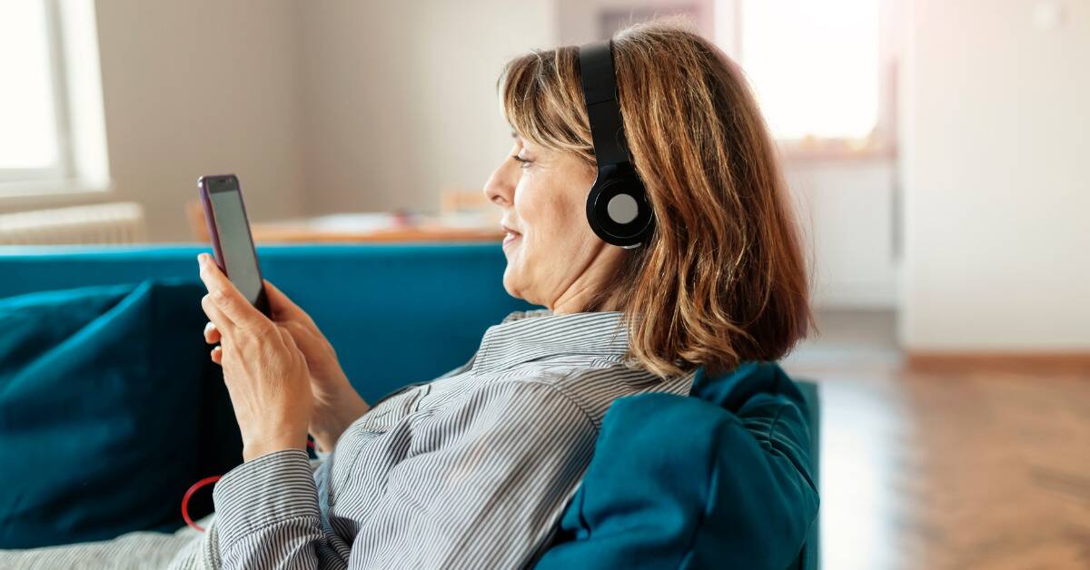 A woman sitting on a couch with headphones on, looking at her phone.