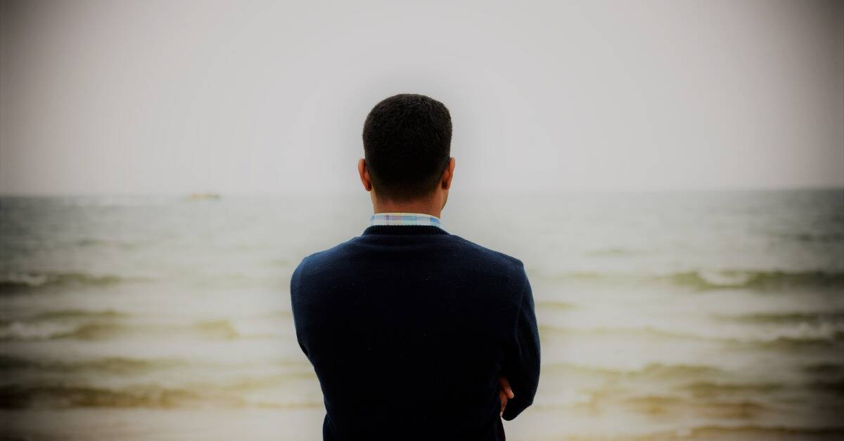 A man with his arms crossed facing the water, a black vignette effect around the edges.