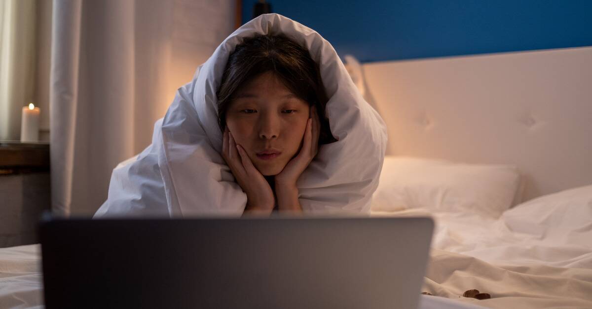 A girl wrapped in a blanket, laying on her bed, watching something on her laptop.