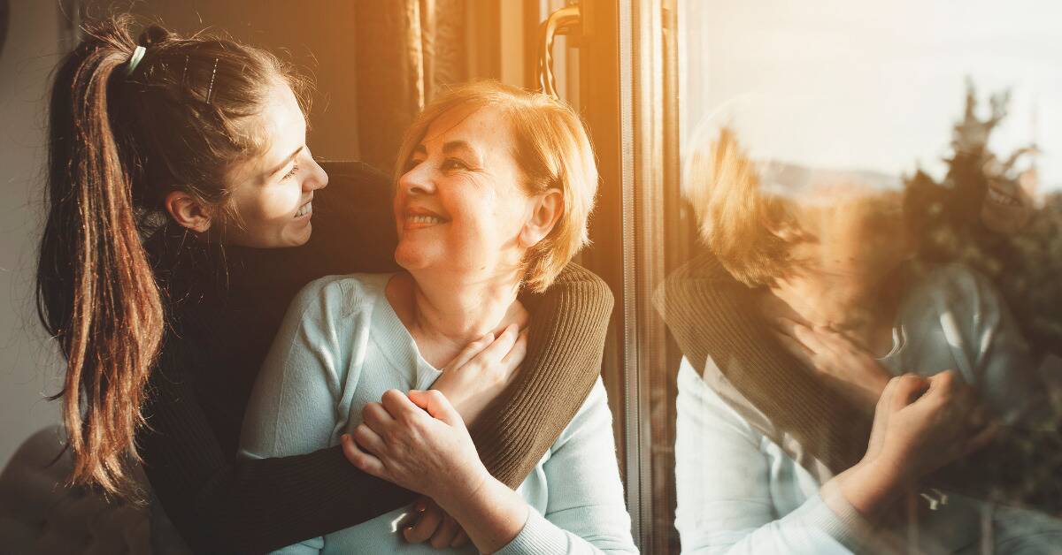 Two women of different generations hugging, smiling at one another, next to a sunny window.