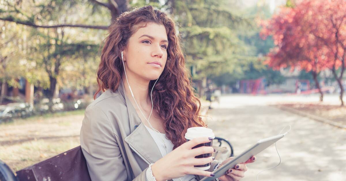 A woman sitting on a bench, holding an iPad and a coffee cup, looking thoughtful.
