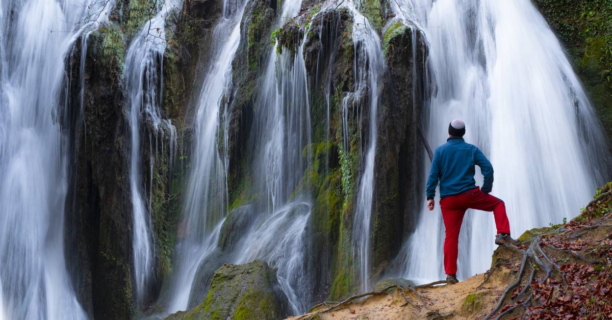 A man during a hike, having stopped to look at a collection of waterfalls, leg propped up on a ledge.