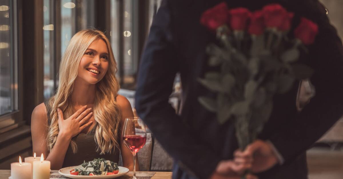 A woman sitting at a table for a date, the man holding a bouquet of roses behind his back.