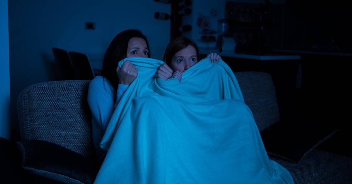 Two women watching a horror movie, lit in blue, a blanket pulled up to their eyes.