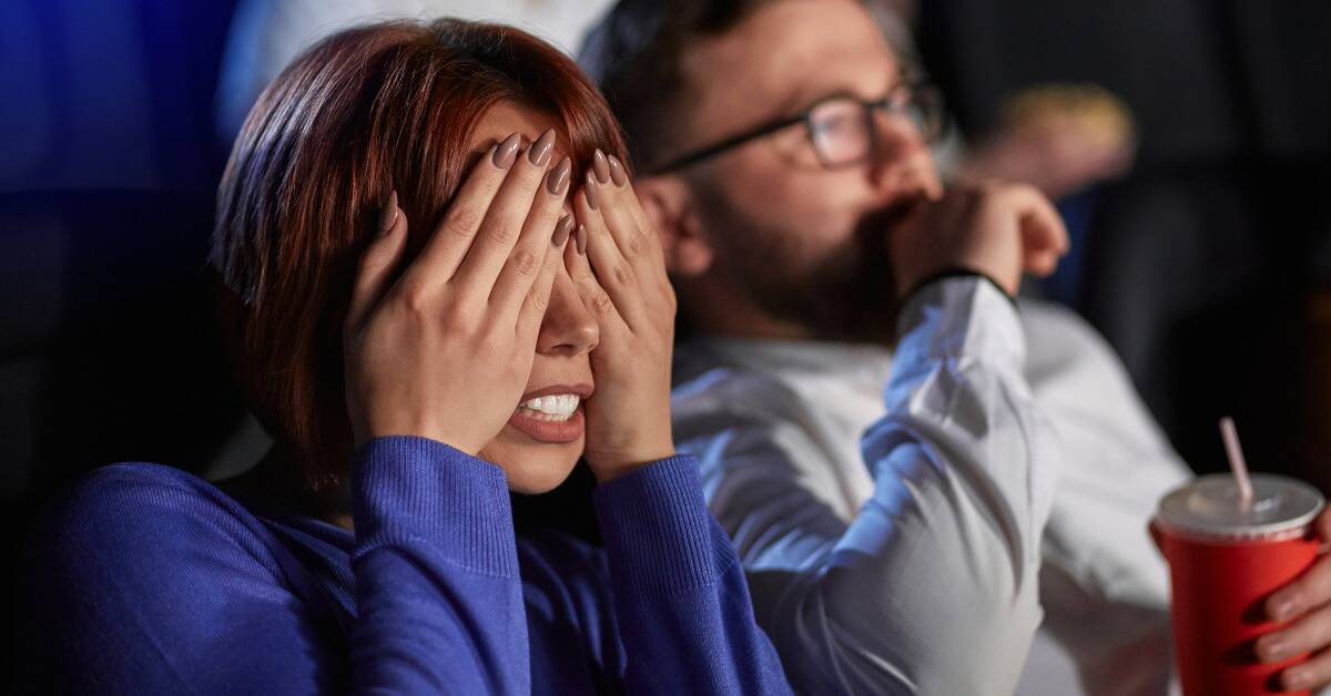 Two people in a movie theater watching a horror movie, the woman covering her eyes with her hands.