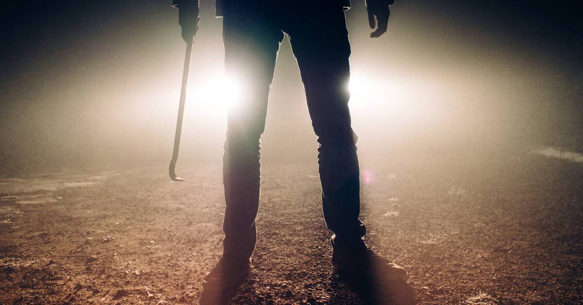 The lower half silhouette of someone standing in front of car headlights, holding a crowbar in one hand.