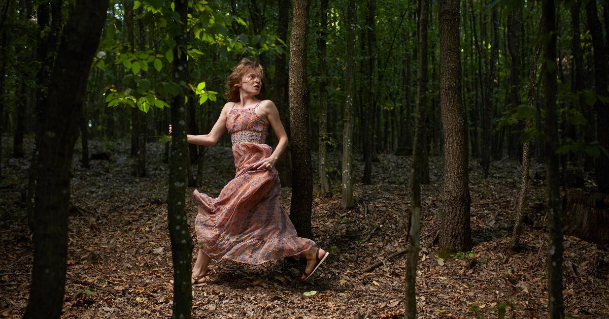 A woman running through the forest in a pink dress, looking over her shoulder.