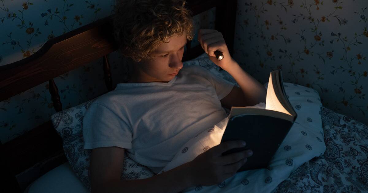 A young boy reading a book in bed via flashlight.