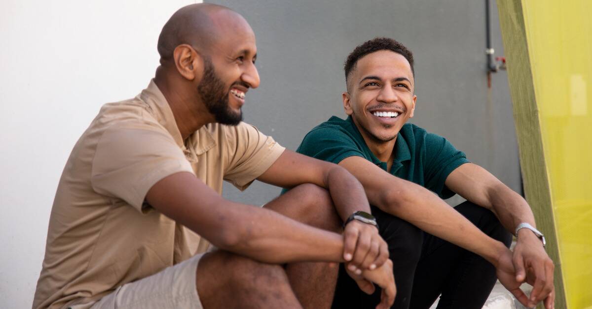 Two male friends sitting side by side and smiling as they talk.