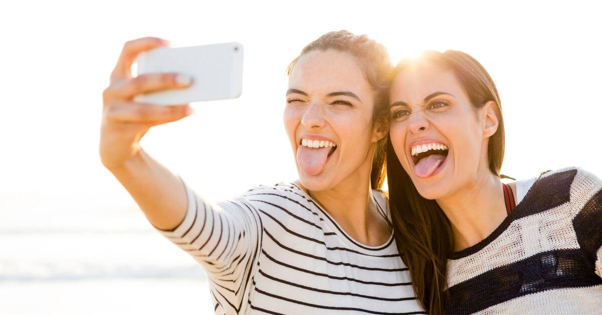 Two female friends sticking their tongues out and making silly faces as they take a selfie.