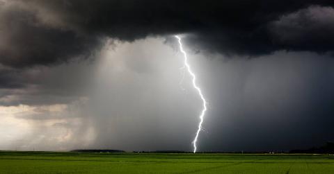 Dark clouds with a single bolt of lightning striking from them onto a crop field, rain pouring behind it.