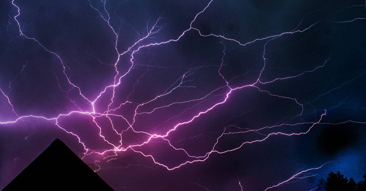 Streaks of pink and purple tinted lightning striking from behind the roof of a home.