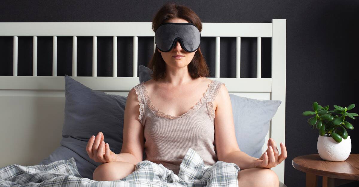 A woman sitting in bed in a meditation post with an eye mask on.