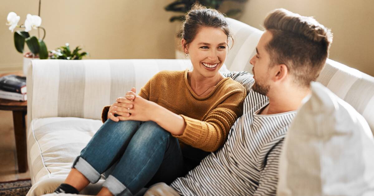 A woman smiling as she looks at her boyfriend, both seated on the couch.