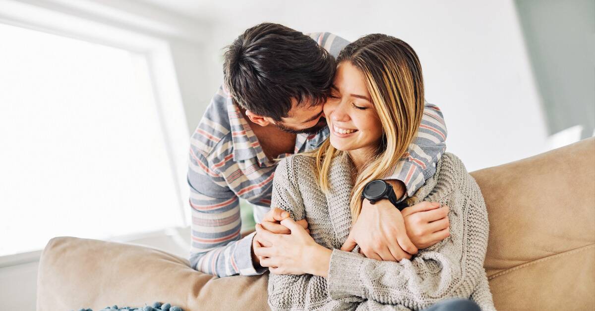 A woman sitting on a couch, smiling as her boyfriend comes up behind her and hugs her.