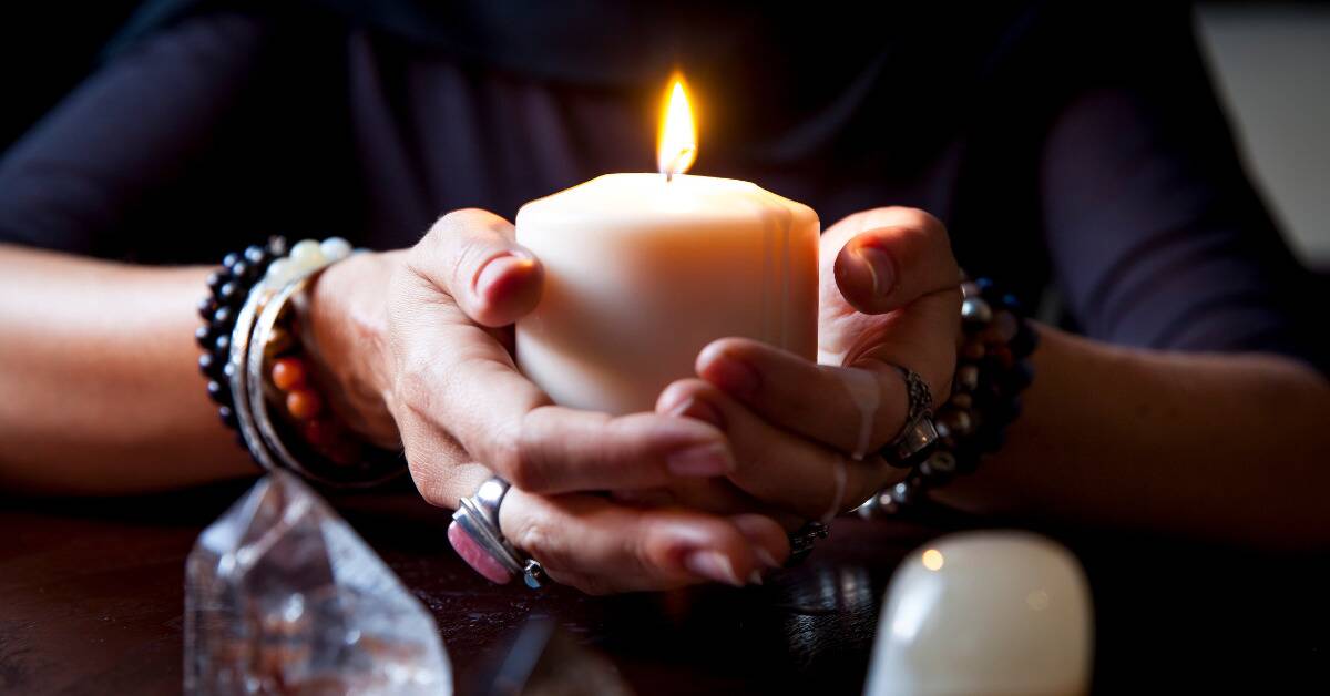 A woman holding a lit candle in her cupped hands, adorned with bracelets and rings.