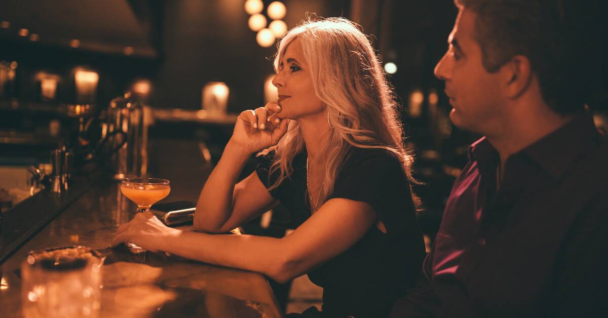 A woman and a man sitting at a bar, the man looking at the woman, the woman looking forward, hand on her drink.