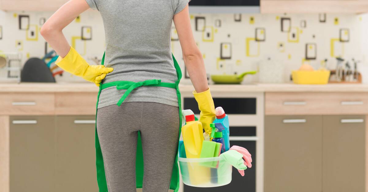 A woman standing in front of her kitchen, armed with a bucket of leaning supplies.