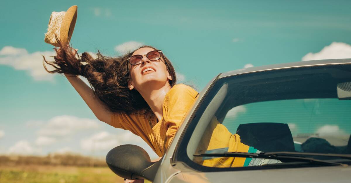 A woman smiling as she leans out a car window, her hair blowing in the wind.
