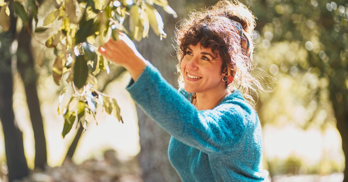 A woman outside, smiling as she picks something off a tree branch.