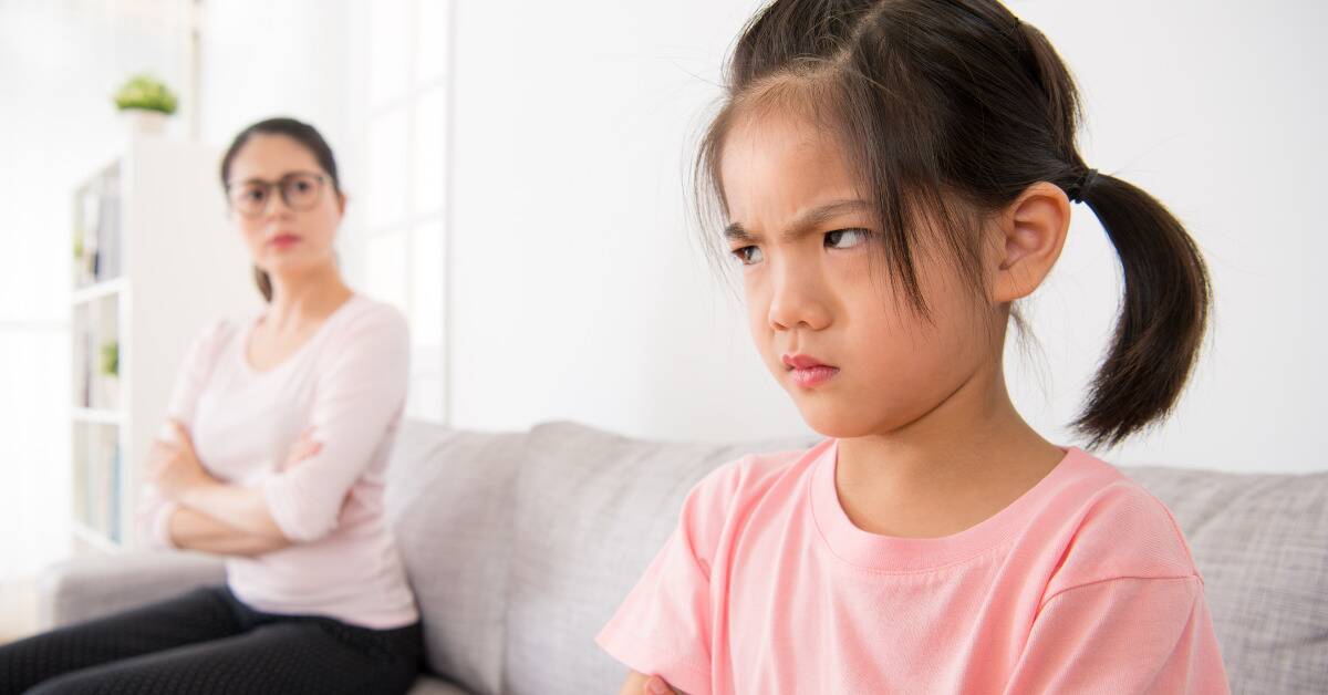 A young girl looking angry, side-eyeing her mother who's sat off to the side with her arms crossed.