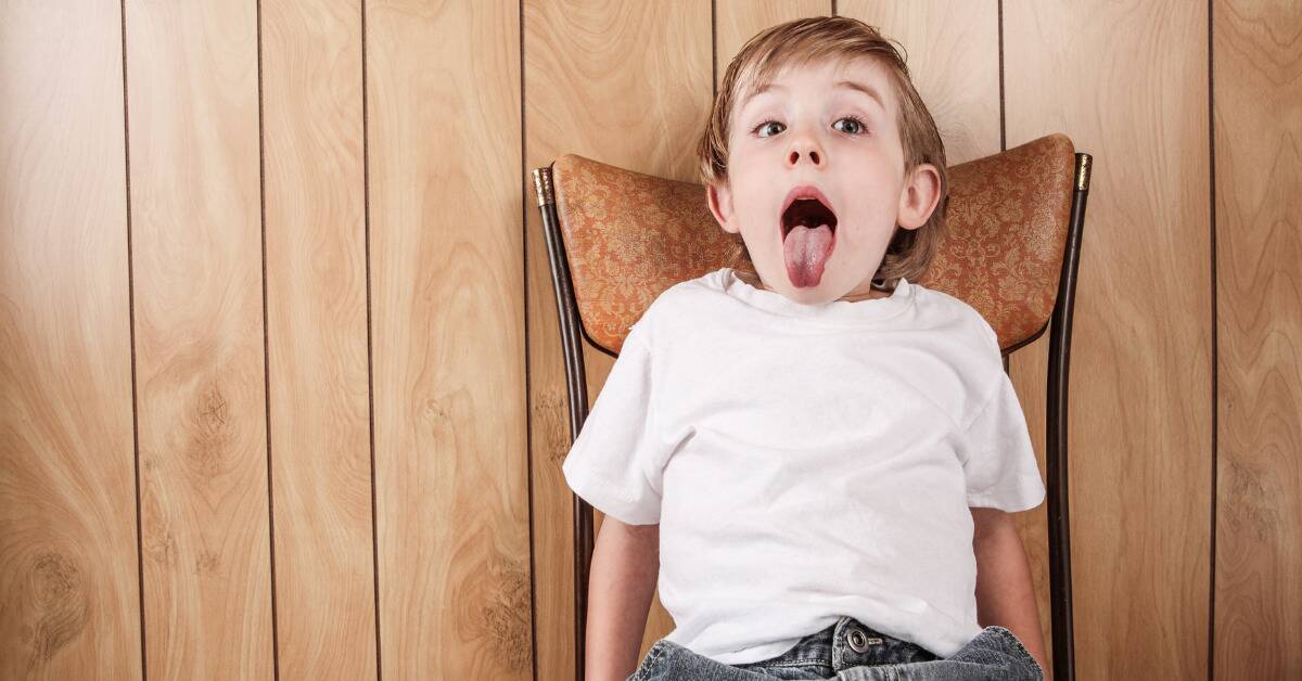 A rambunctious kid sitting on a chair, sticking his tongue out.