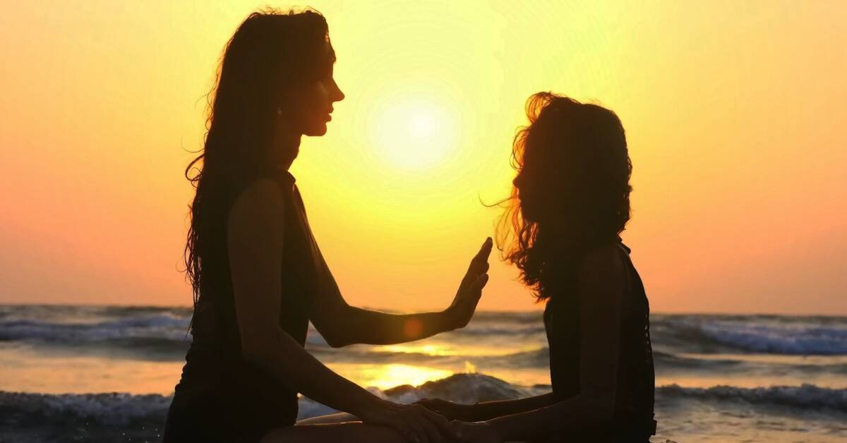 A woman engaging in some spiritual healing with her daughter, both sat on a beach at sunset.
