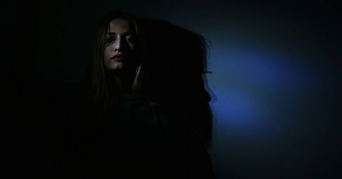 A woman mostly shrouded in darkness, her face only slightly visible in shadow.