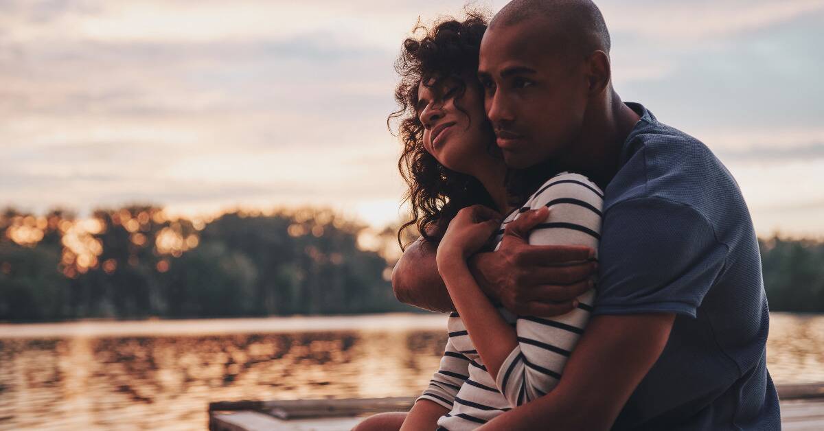 A couple is sat outside, the man wrapping his arms around the woman from behind.