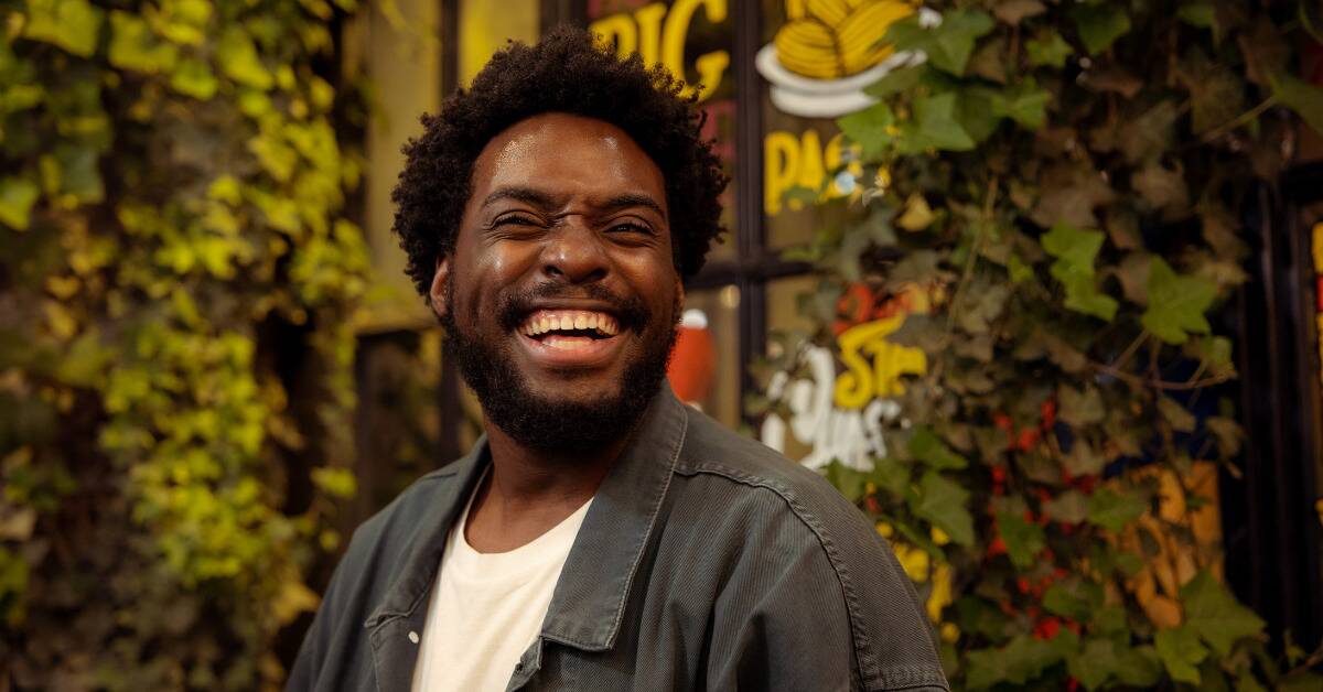 A man standing in front of a storefront covered in vines smiles brightly.