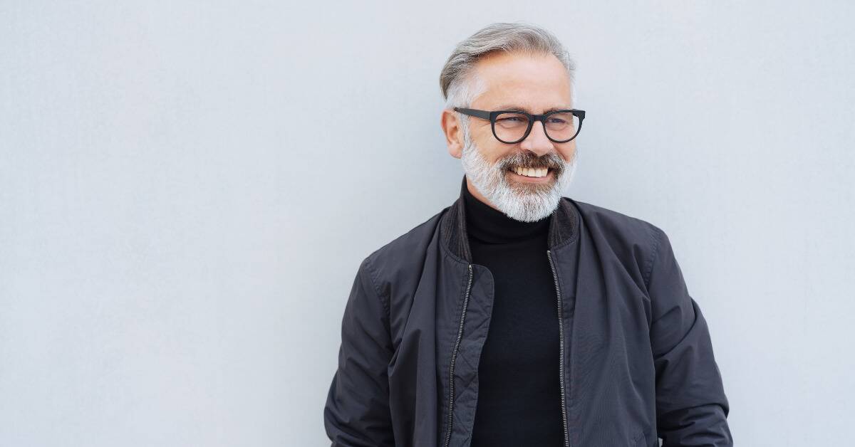 An older, stylish man smiles as he looks to the sit and leans against a white wall.