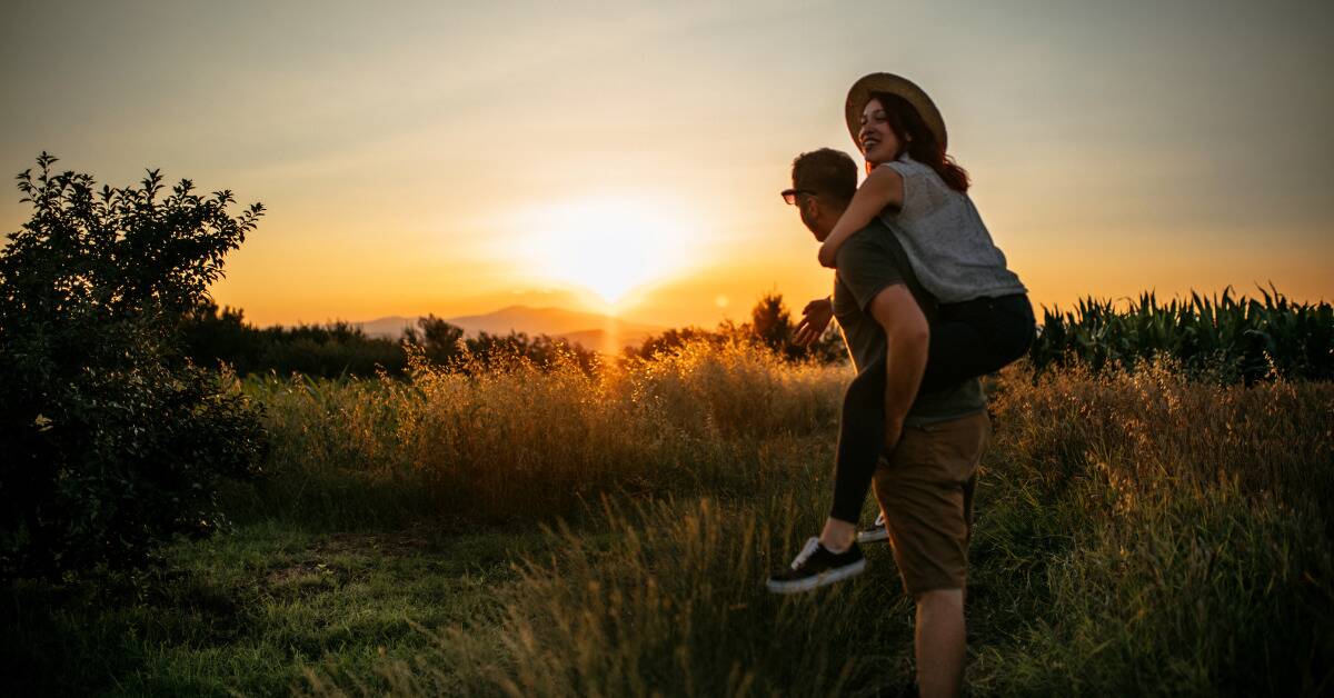 A couple out walking in a field at sunset, the man giving the woman a piggyback ride.