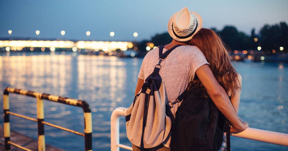 A couple standing on a pier, looking out at the water, the man's arm around the woman.