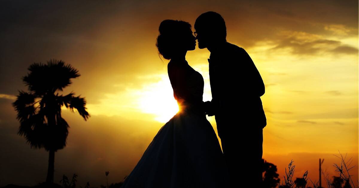 A silhouette of a couple in front of a sunset. The woman is in a ballgown-esque dress, her hair done up, and the man is leaning in to kiss her.