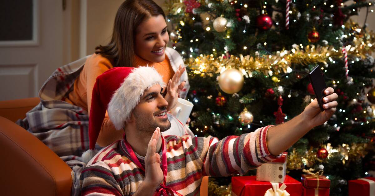 A man and a woman waving into the man's phone, a Christmas setup behind them.
