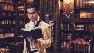 A man in a very fancy suit reading an old book in an old library.