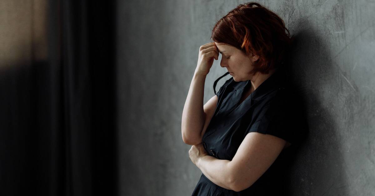 A woman leaning against a wall, hand to her forehead, looking dejected.