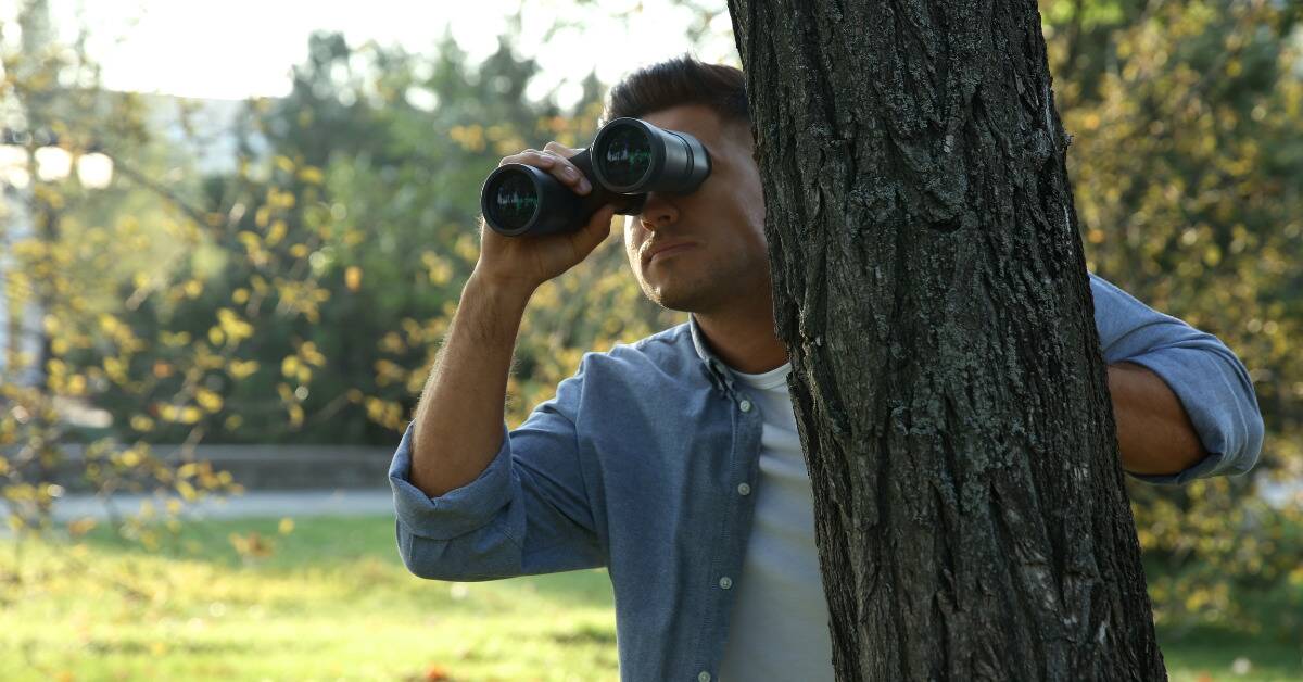A man behind a tree looking out with a pair of binoculars.