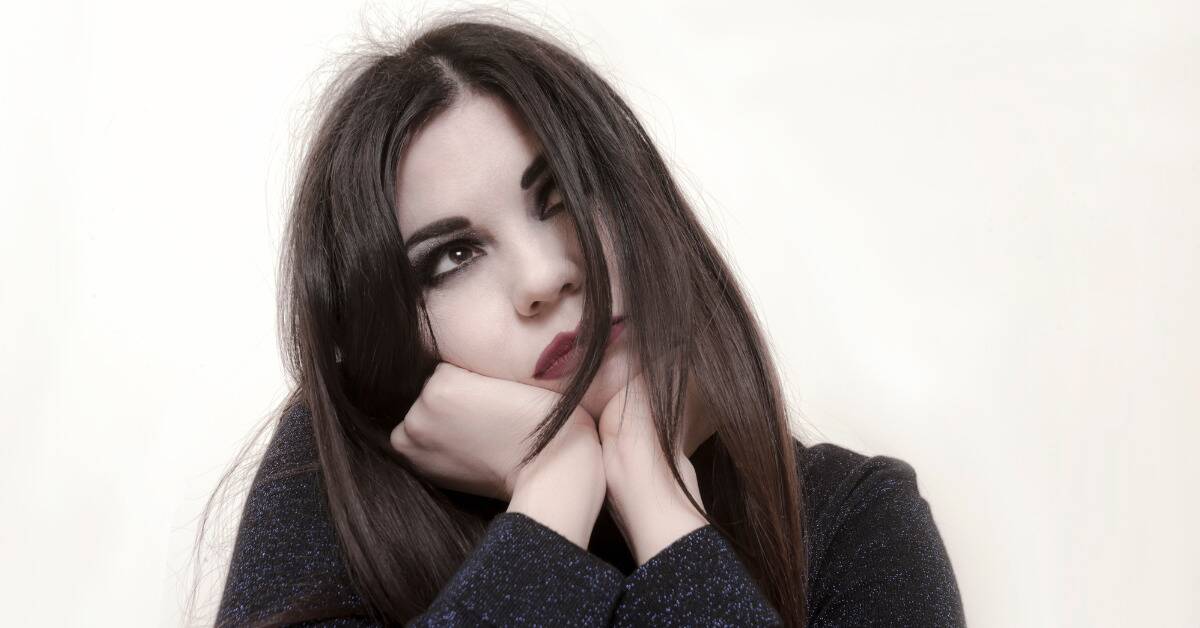 A woman with dark makeup resting her chin on her hands and looking wistfully up to the sky.