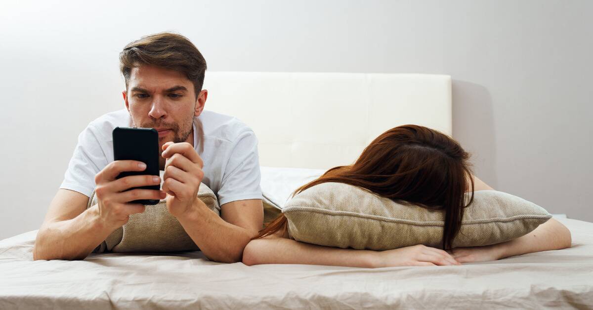 A couple in bed, both laying on their stomachs, the woman asleep while the man uses his phone.