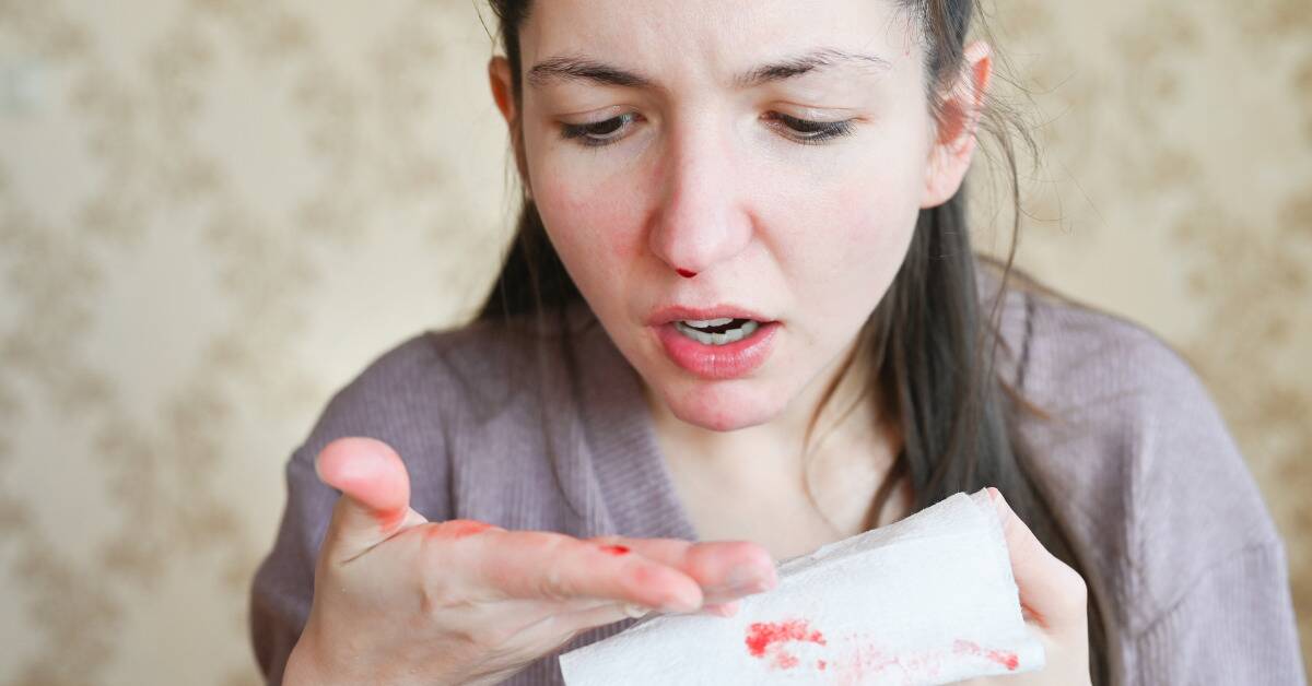 A woman looking down at her hand in shock, revealing blood that came from her nose.