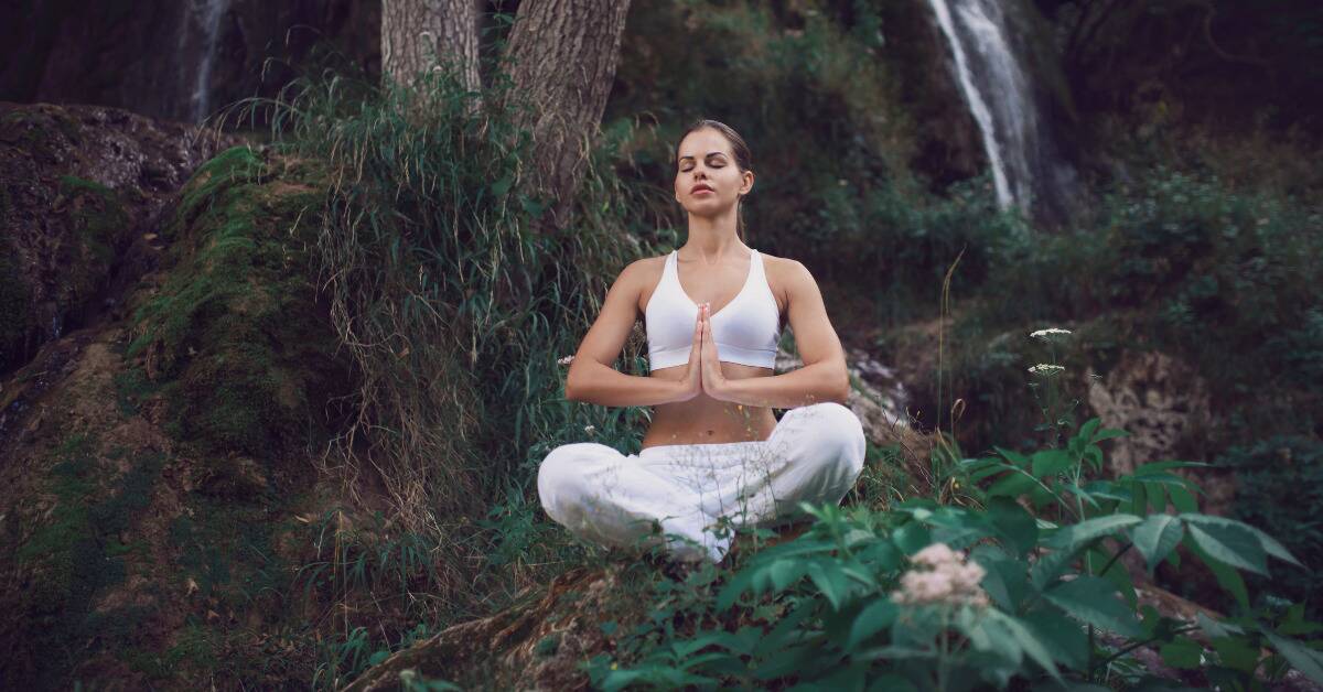 A woman meditating in nature, legs crossed and hands together.