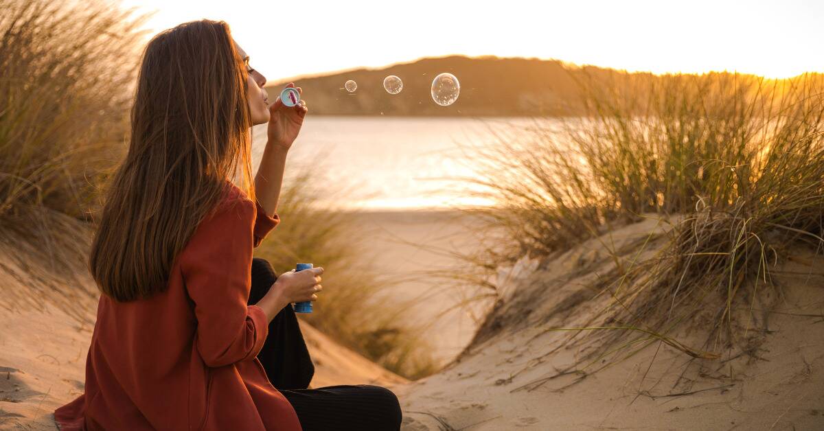 A woman sitting on a beach blowing bubbles.